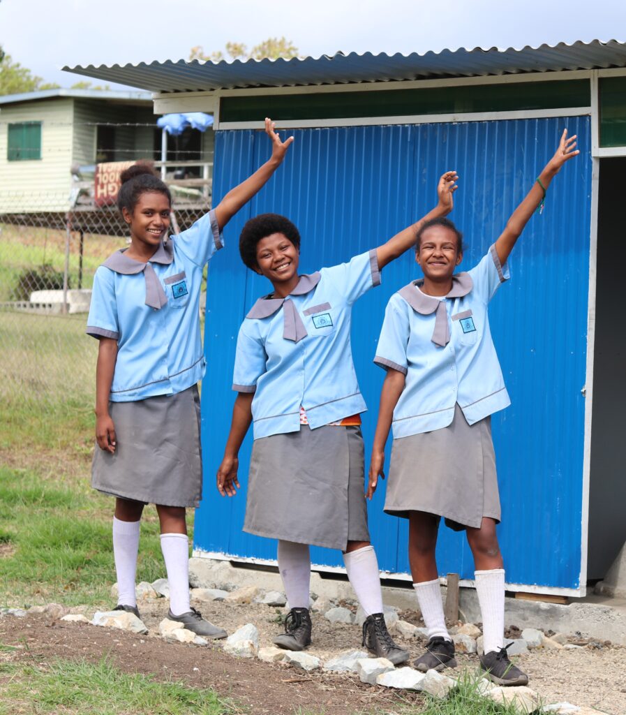 Adolescent girls in blue school uniforms outside a blue shelter with their hands raised.