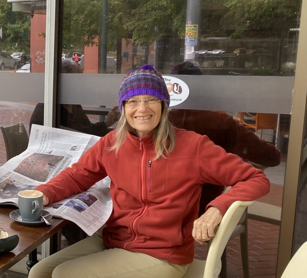 A woman in a red fleece jacket and a purple beanie sits at a table with a coffee and newspaper. She is turned toward the camera and smiling.