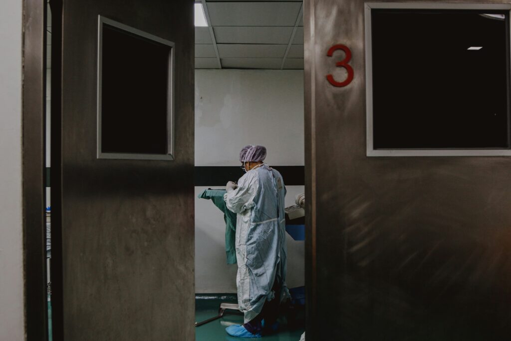 A doctor wearing scrubs stands behind an open hospital door, facing away from the camera. Photo by SJ Objio on Unsplash