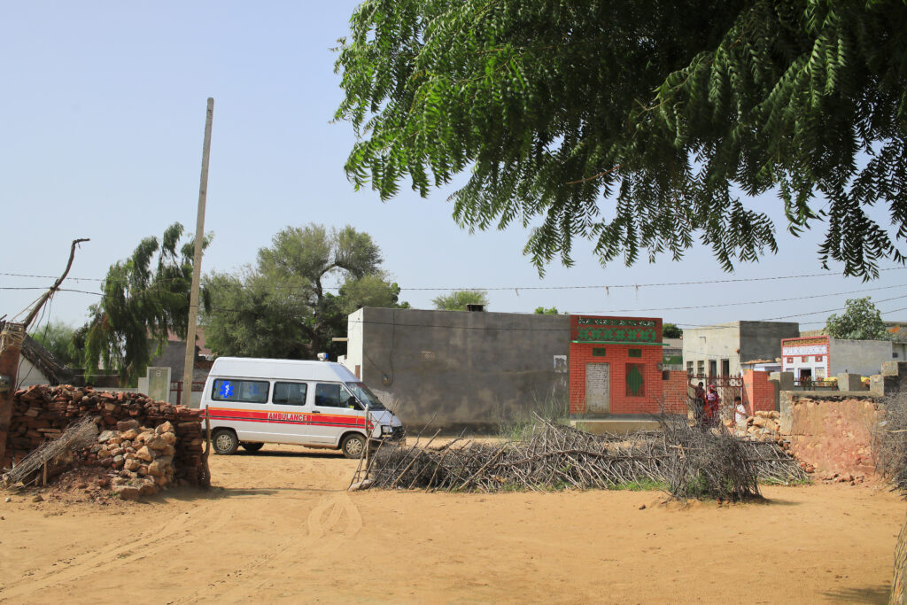 A white and orange van drives through a village in India. A tree hangs overhead, and there are buildings in the background. The sun is shining.