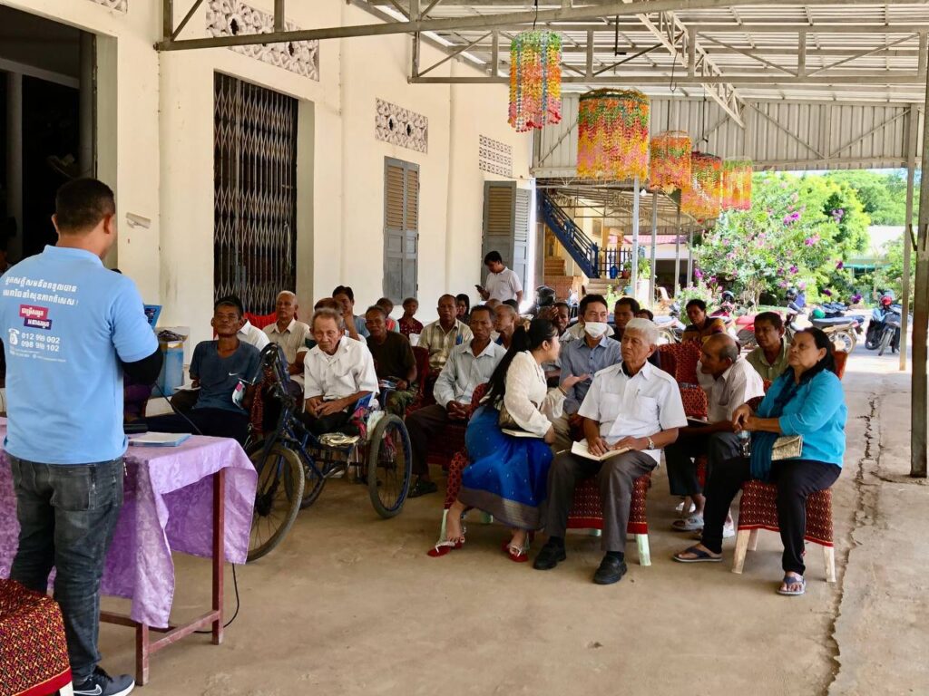 A man in a blue shirt presents to a group of people at an information session for people with disabilities in Cambodia. They are seated outside a white building.