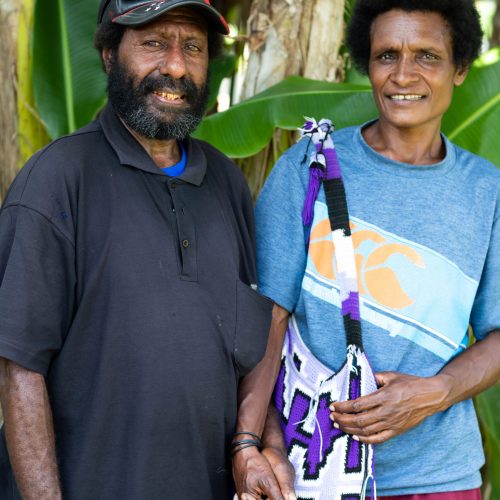 A man and woman look at the camera smiling. They are holding hands, and she is carrying a woven bag on her shoulder.